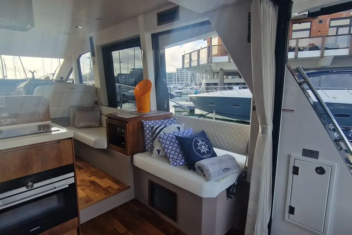Interior of a yacht featuring a kitchen area, cushioned seating with nautical-themed pillows, and a view of moored boats.